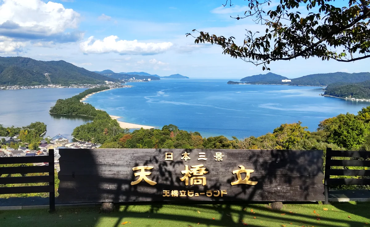 Amanohashidate sandbar seen beyond a wooden lookout sign under a blue sky with scattered clouds