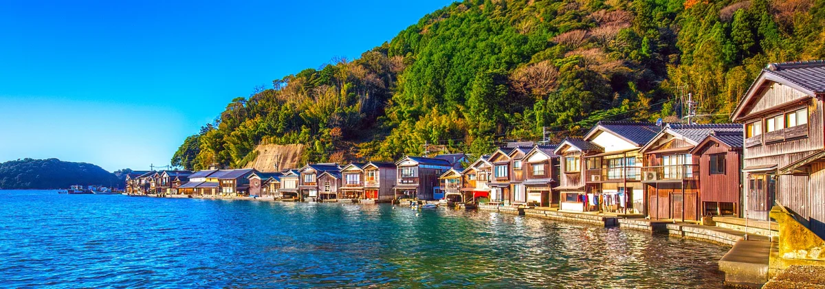 A long row of wooden funaya boathouses beneath wooded hills beside blue water in Ine