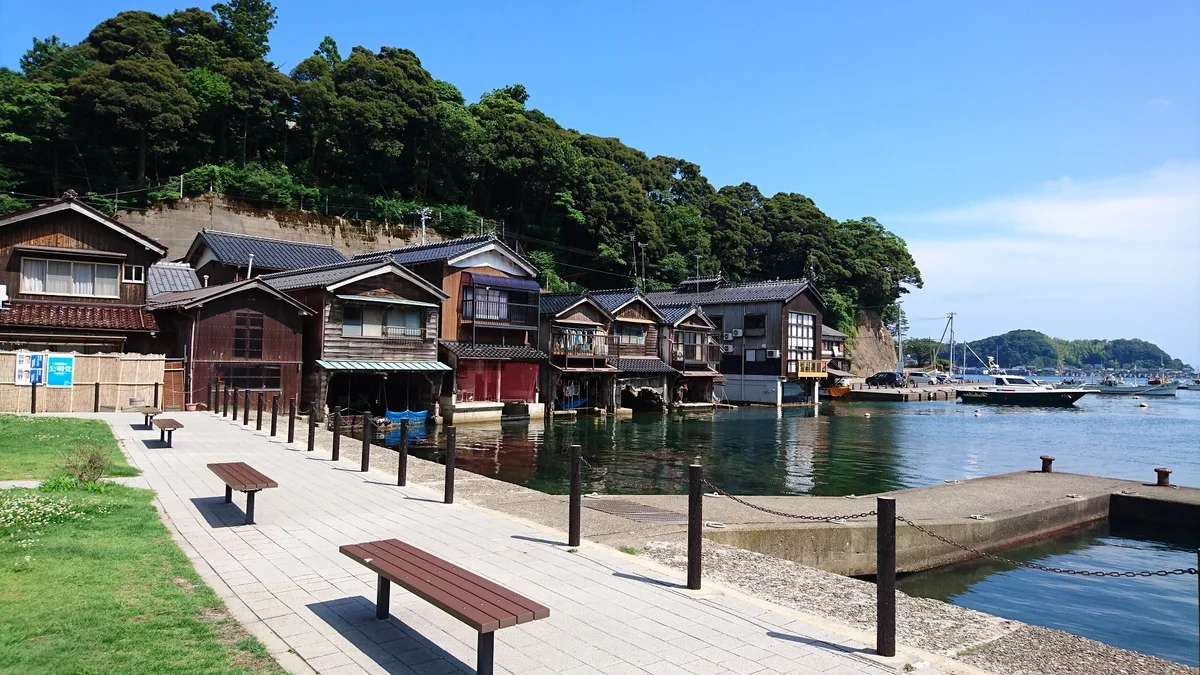 Waterfront benches and a paved walkway beside traditional wooden boathouses in Ine