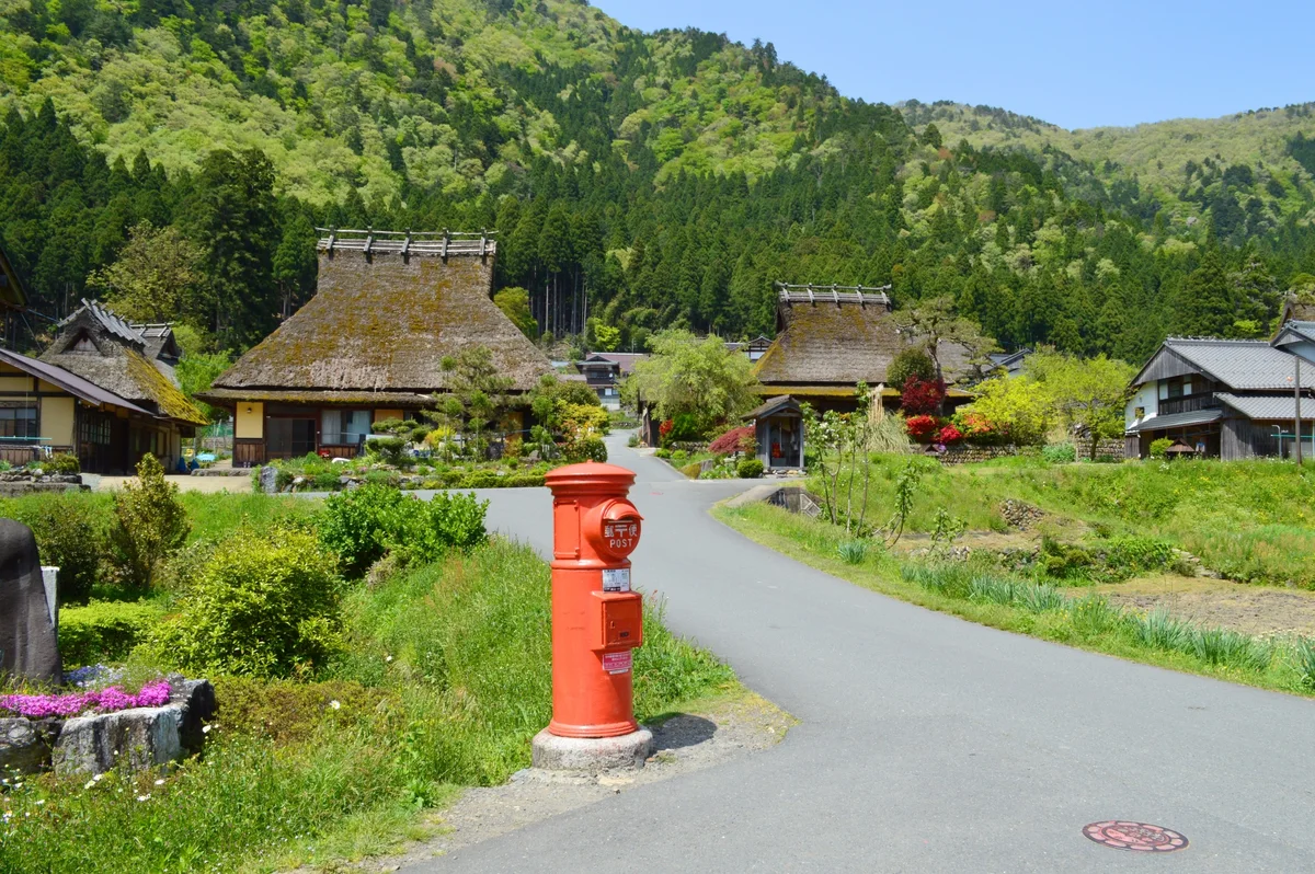 A red postbox beside a village lane and thatched-roof houses backed by green hills in Miyama