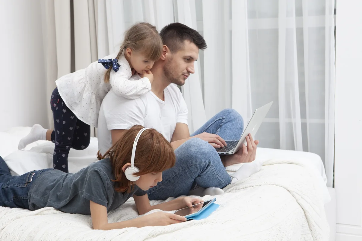 A parent and two children using a laptop and tablet together on a bed