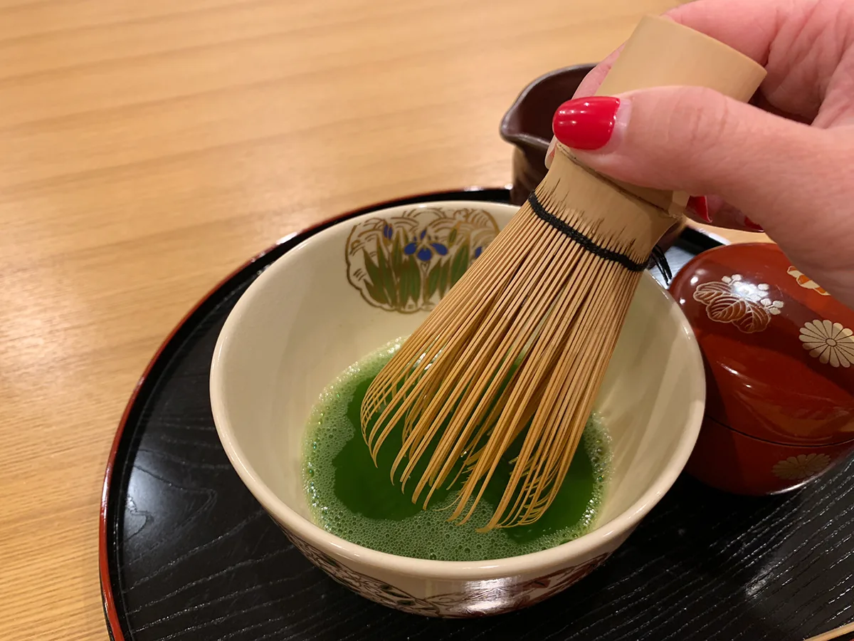 A hand using a bamboo whisk to prepare matcha in a ceramic bowl