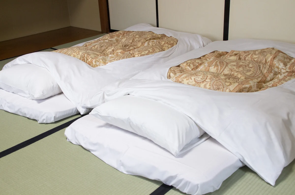 Two futons with white bedding and patterned quilts laid side by side on tatami flooring