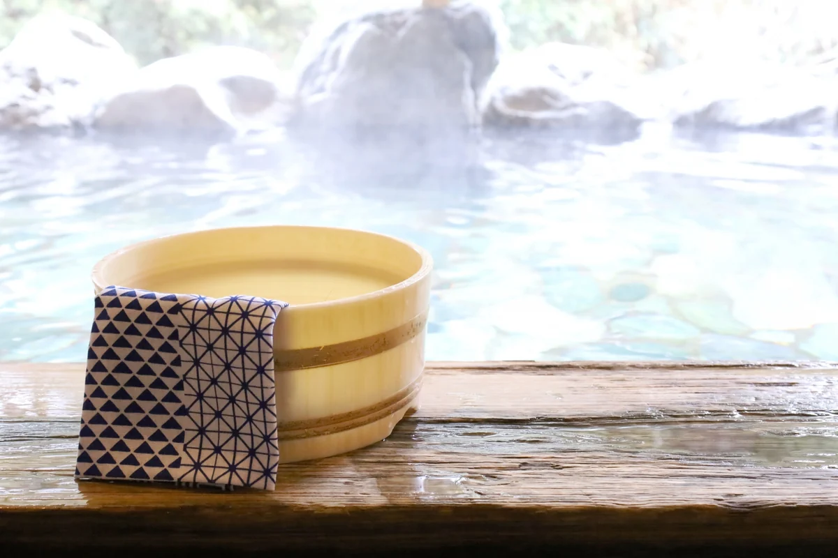 Wooden wash bucket and patterned towel beside a steaming outdoor bath bordered by rocks