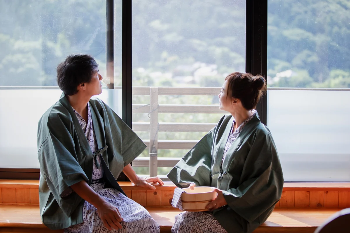 Two guests wearing patterned yukata and green outer jackets, seated beside a window with a wooden wash bucket