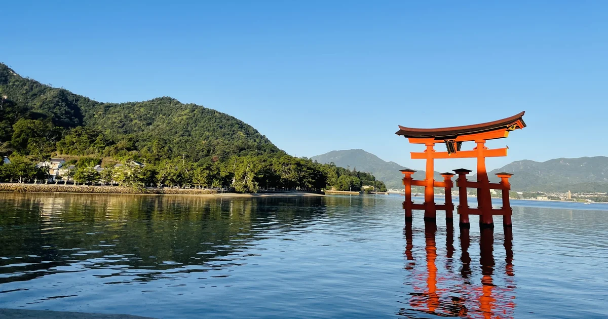 Itsukushima Shrine's vermilion torii reflected in the water beside Miyajima's wooded shore