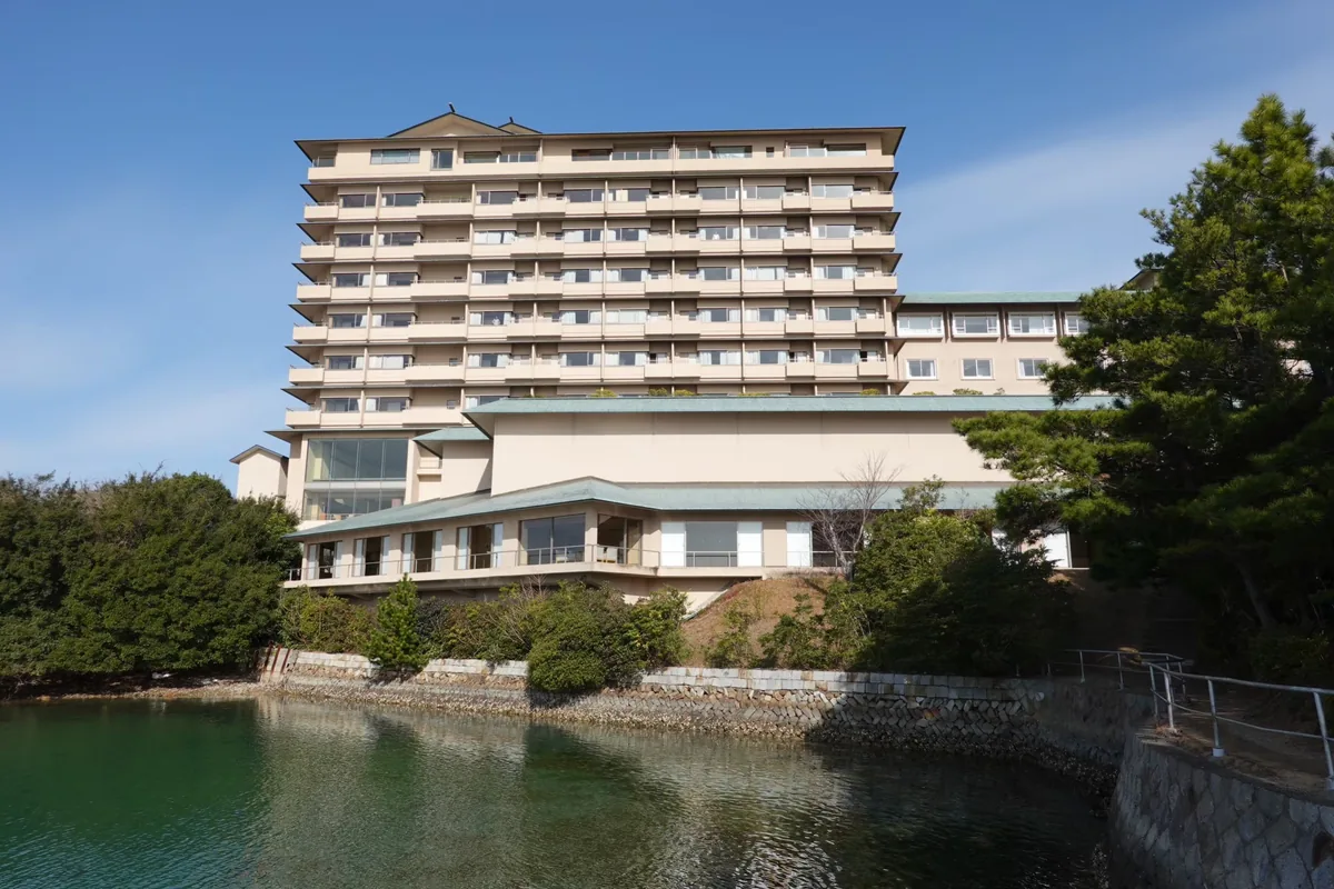 Kashikojima Hojoen rising above a sheltered waterfront, with trees and a stone embankment in the foreground