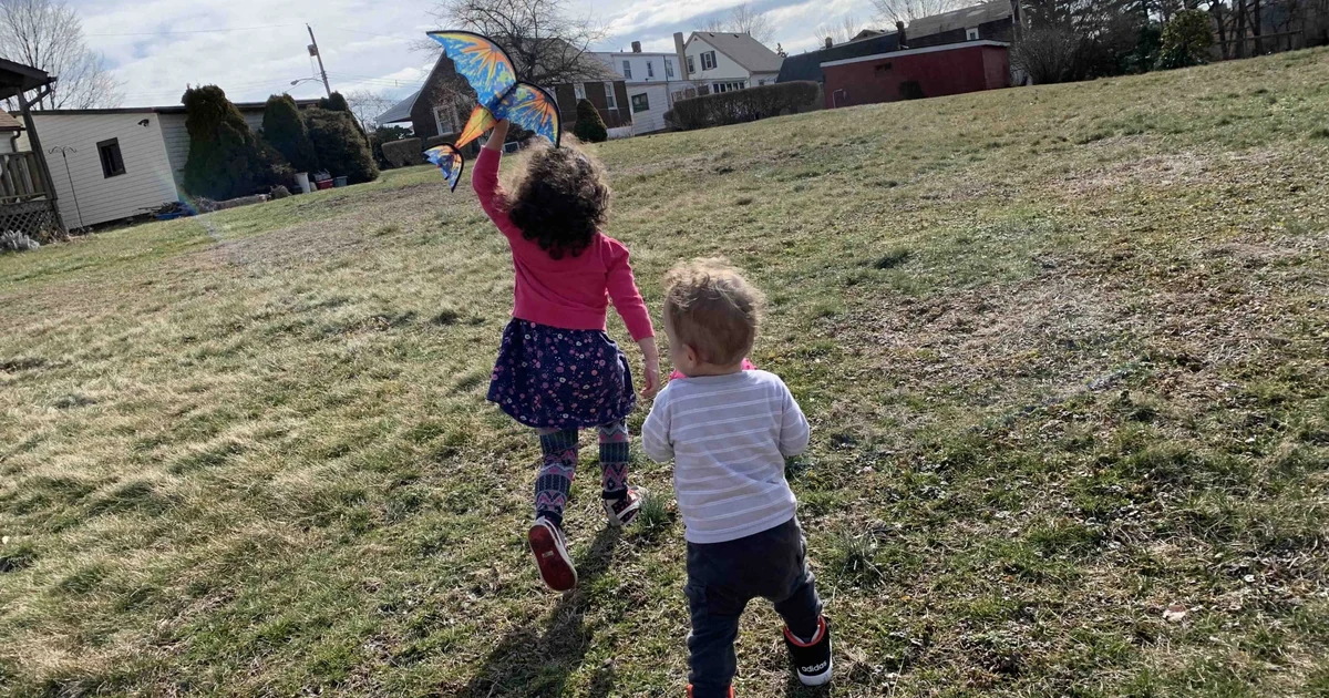 Two children walking across a grassy field, one holding a colorful kite
