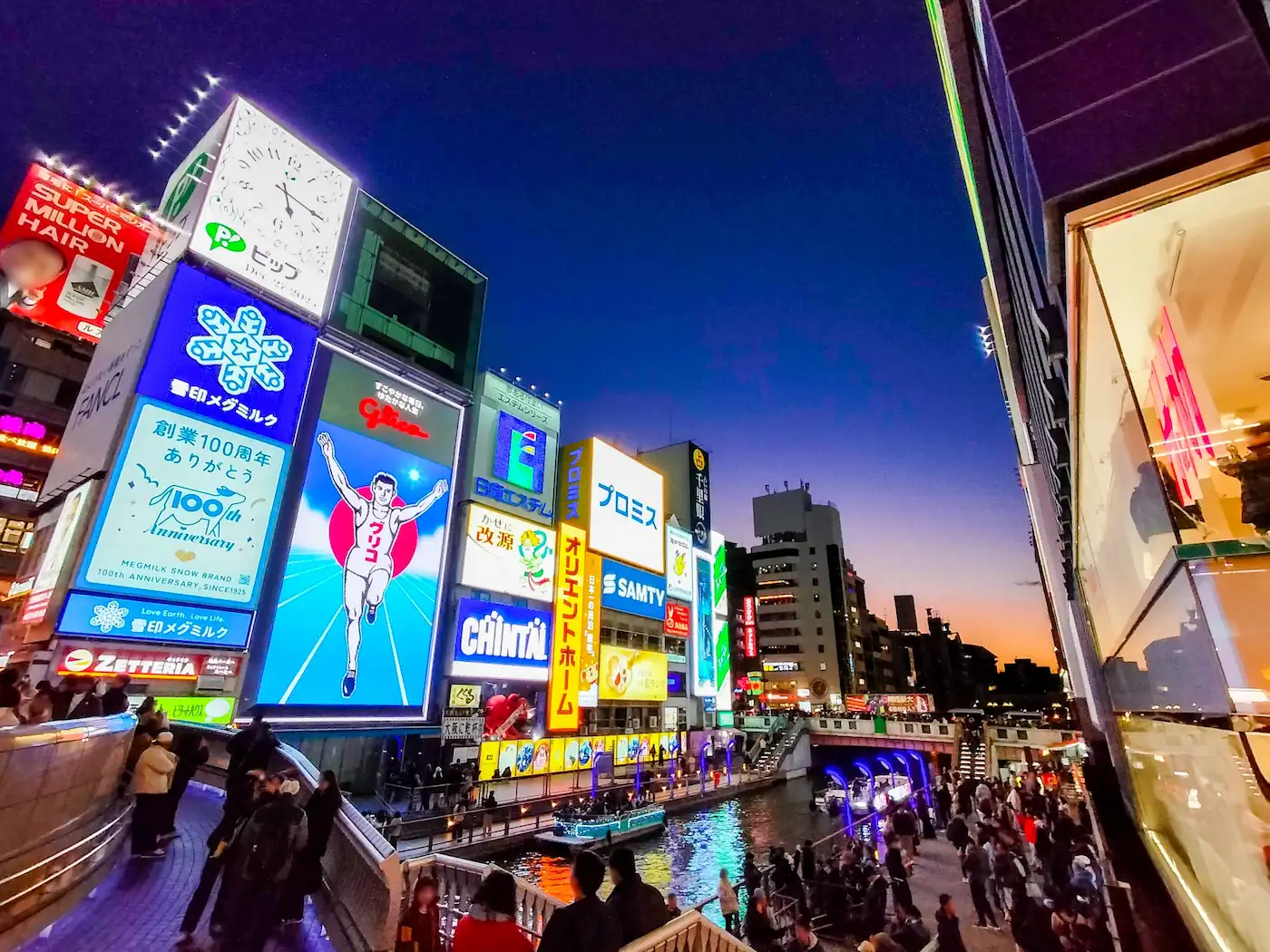 The Dotonbori canal in Osaka at dusk, lit by the Glico running-man sign and neon billboards reflected in the water, with crowds along the walkways.