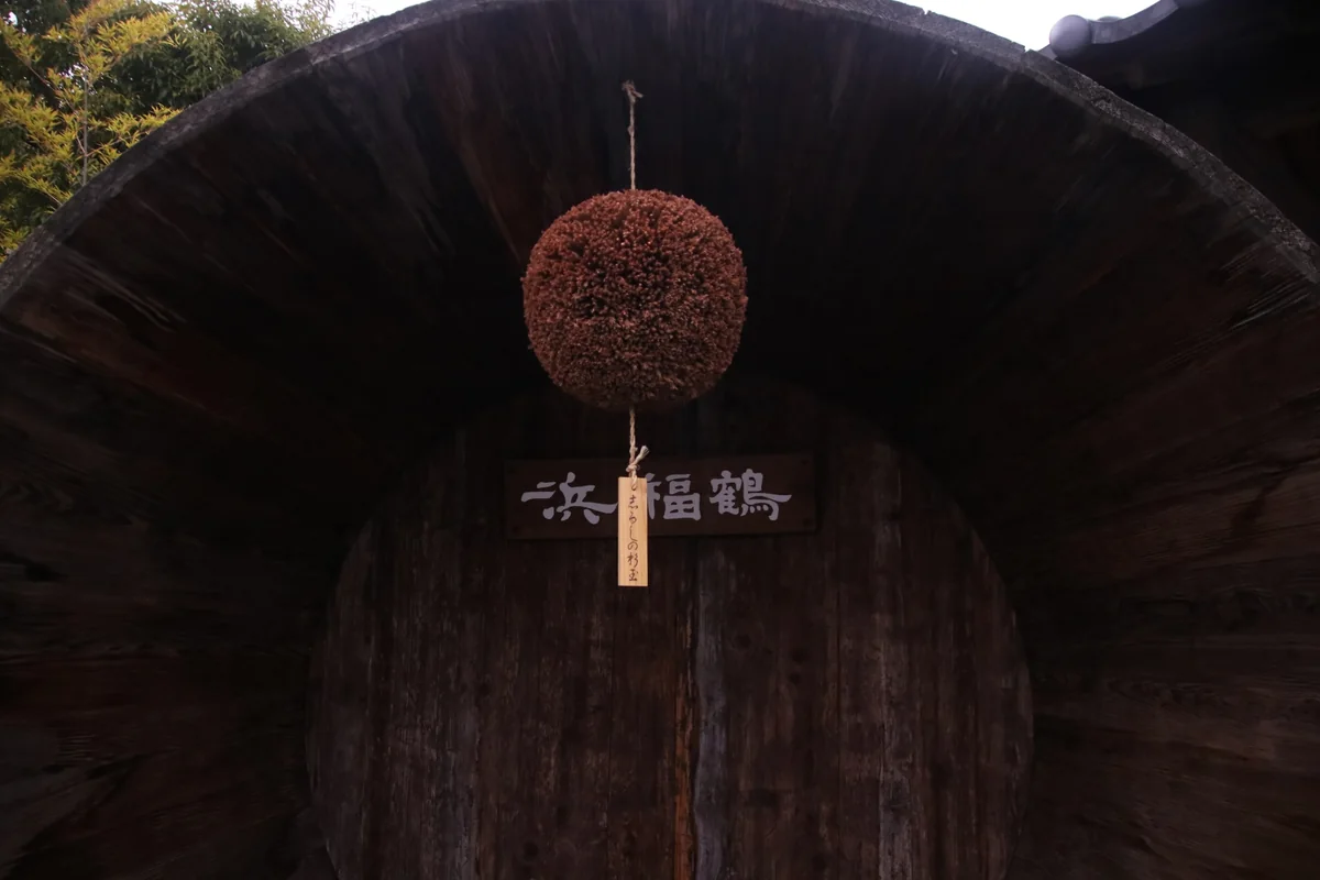A brown sugidama hanging in front of a large wooden sake barrel marked Hamafukutsuru