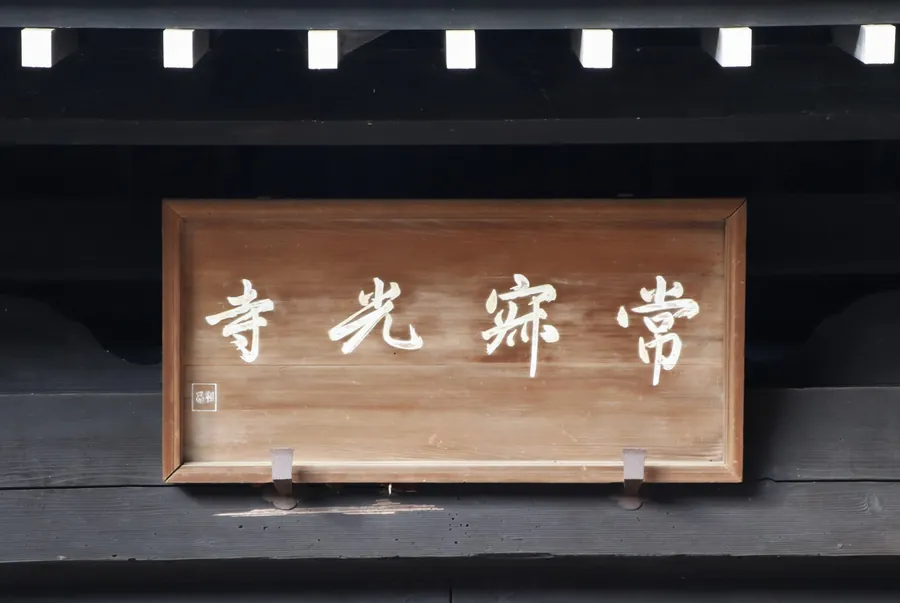 A wooden plaque with the white characters 常寂光寺, read from right to left, mounted on the dark wooden Sanmon gate at Jojakko-ji.