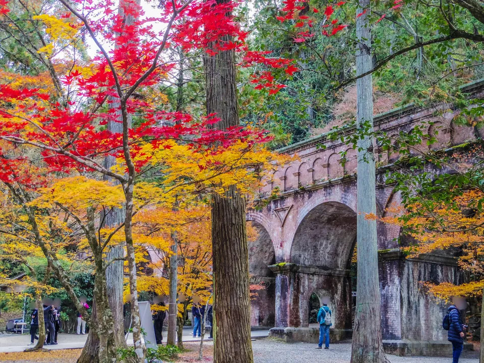 Red and yellow autumn leaves surround the brick Suirokaku aqueduct, with visitors below its arches.