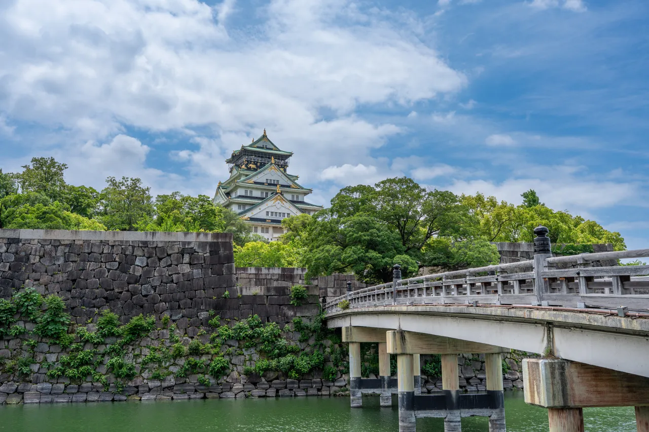 Osaka Castle's main tower above stone walls, green trees and a bridge across the moat