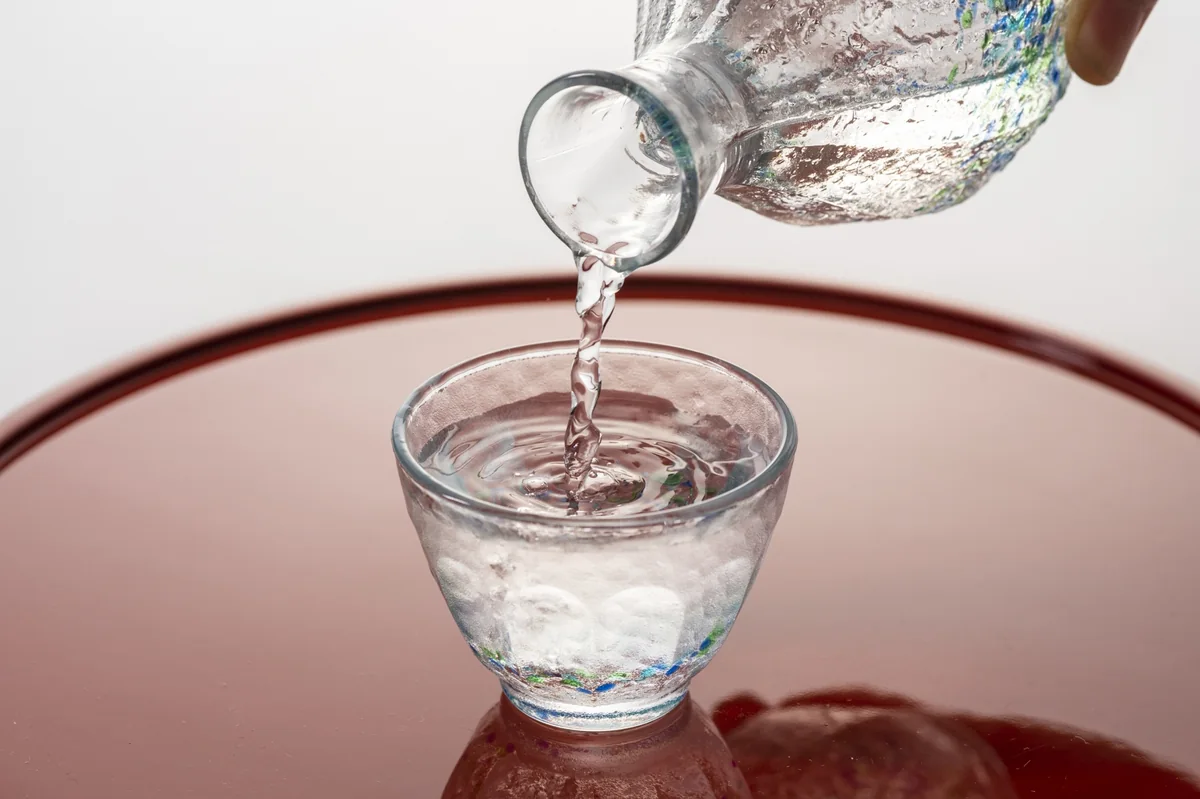 Sake being poured from a glass carafe into a glass cup on a red tray