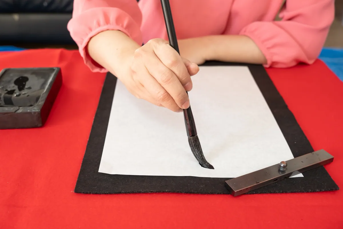 A person in pink sleeves holding a brush over a curved black stroke, with an inkstone beside the paper