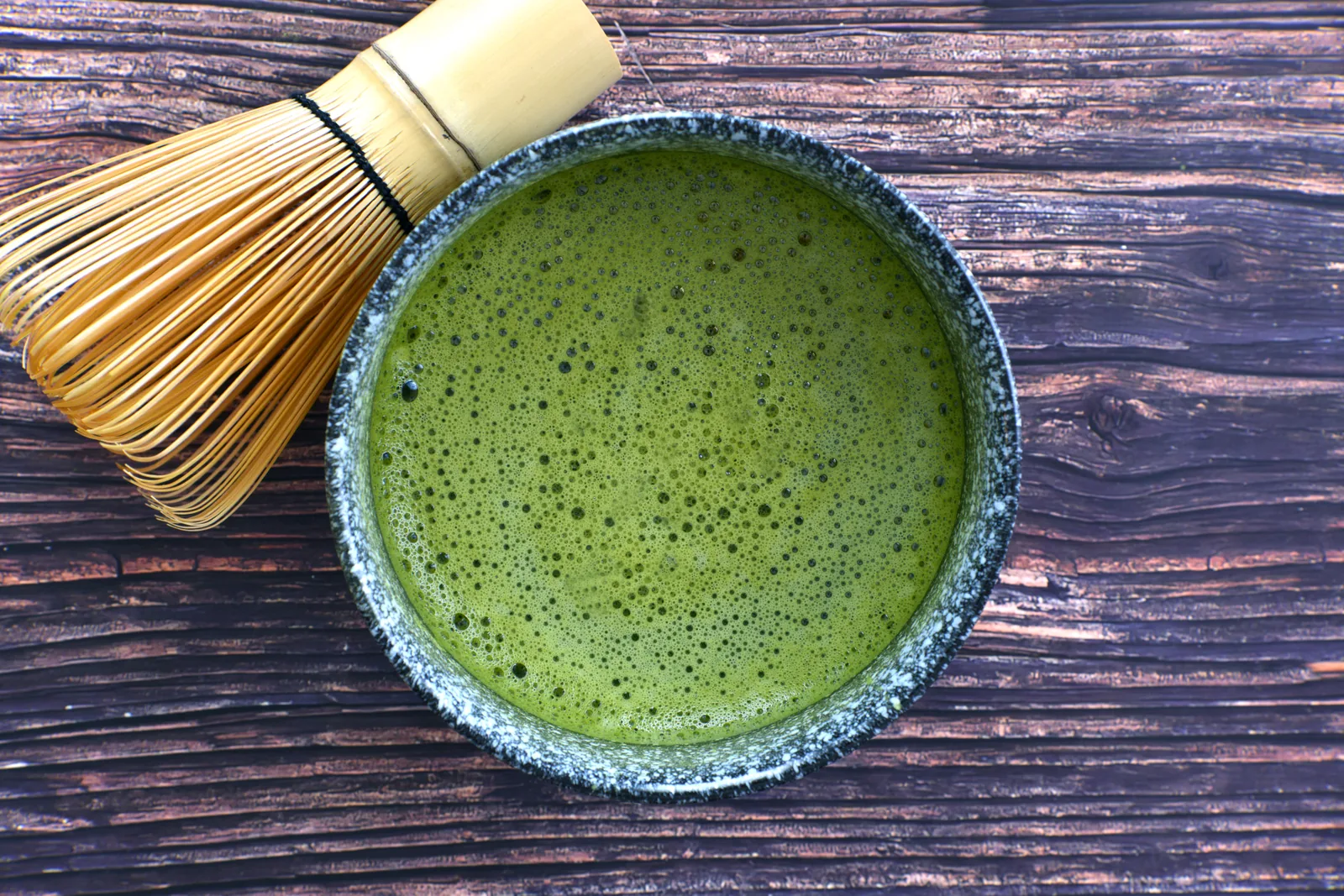 Frothy green matcha in a textured ceramic bowl, with a bamboo whisk on dark wood