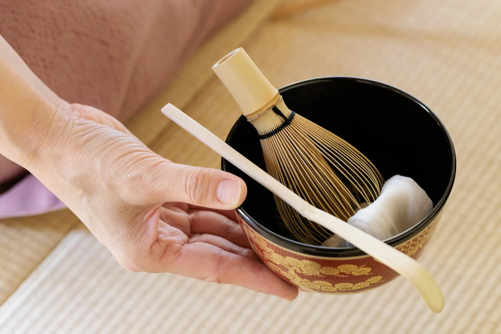 Hands holding a decorated tea bowl containing a bamboo whisk, scoop and cloth, ready for tea preparation