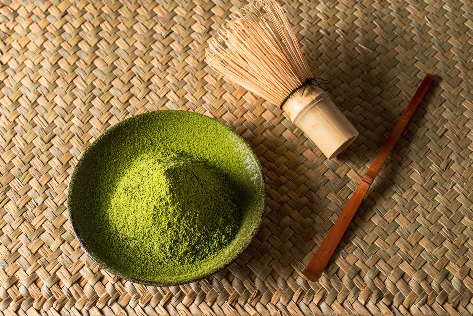 Fine matcha powder in a ceramic dish beside a bamboo whisk and scoop on a woven mat