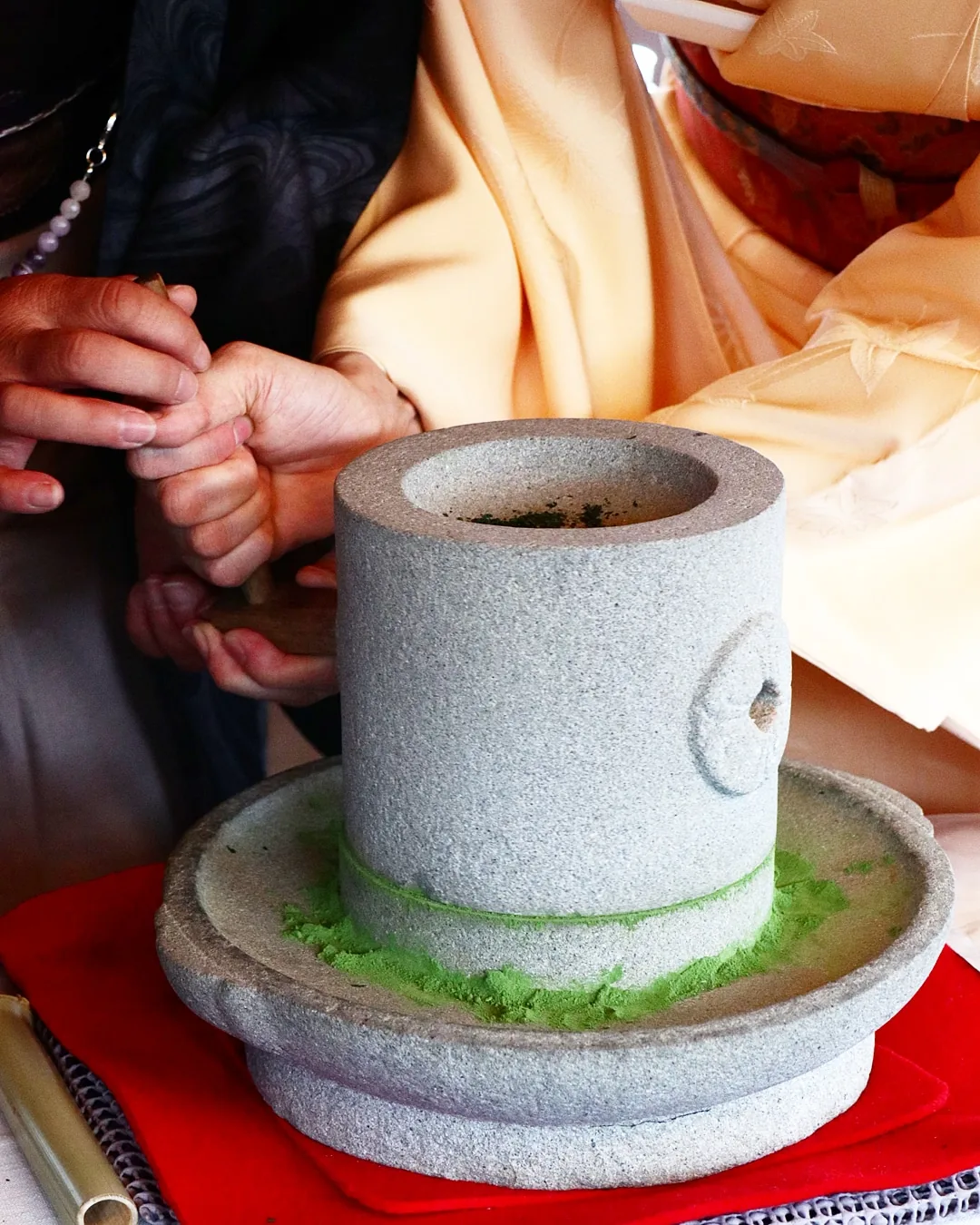One person's hands guiding another's as they turn a stone tea mill, with green powder collecting below