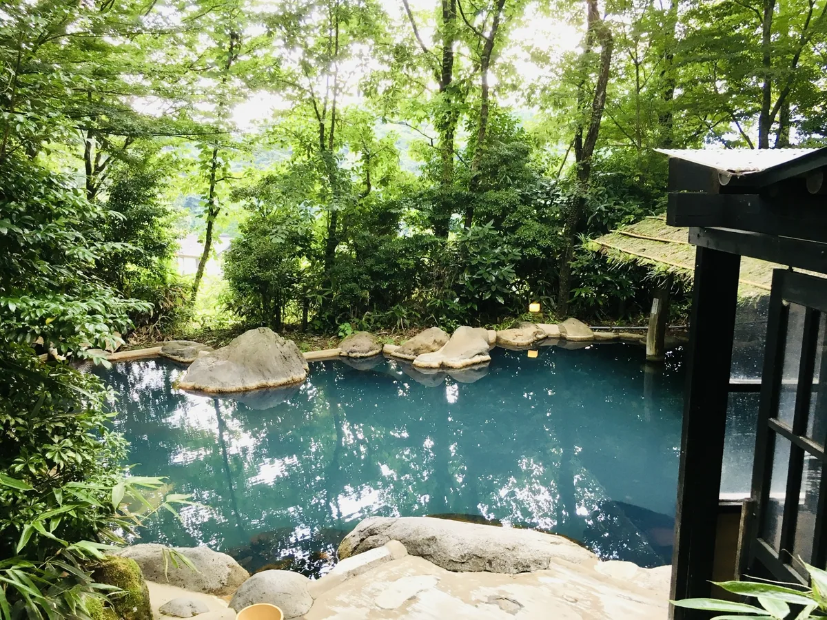 A blue-green outdoor rock bath surrounded by leafy trees, with a wooden shelter at one side