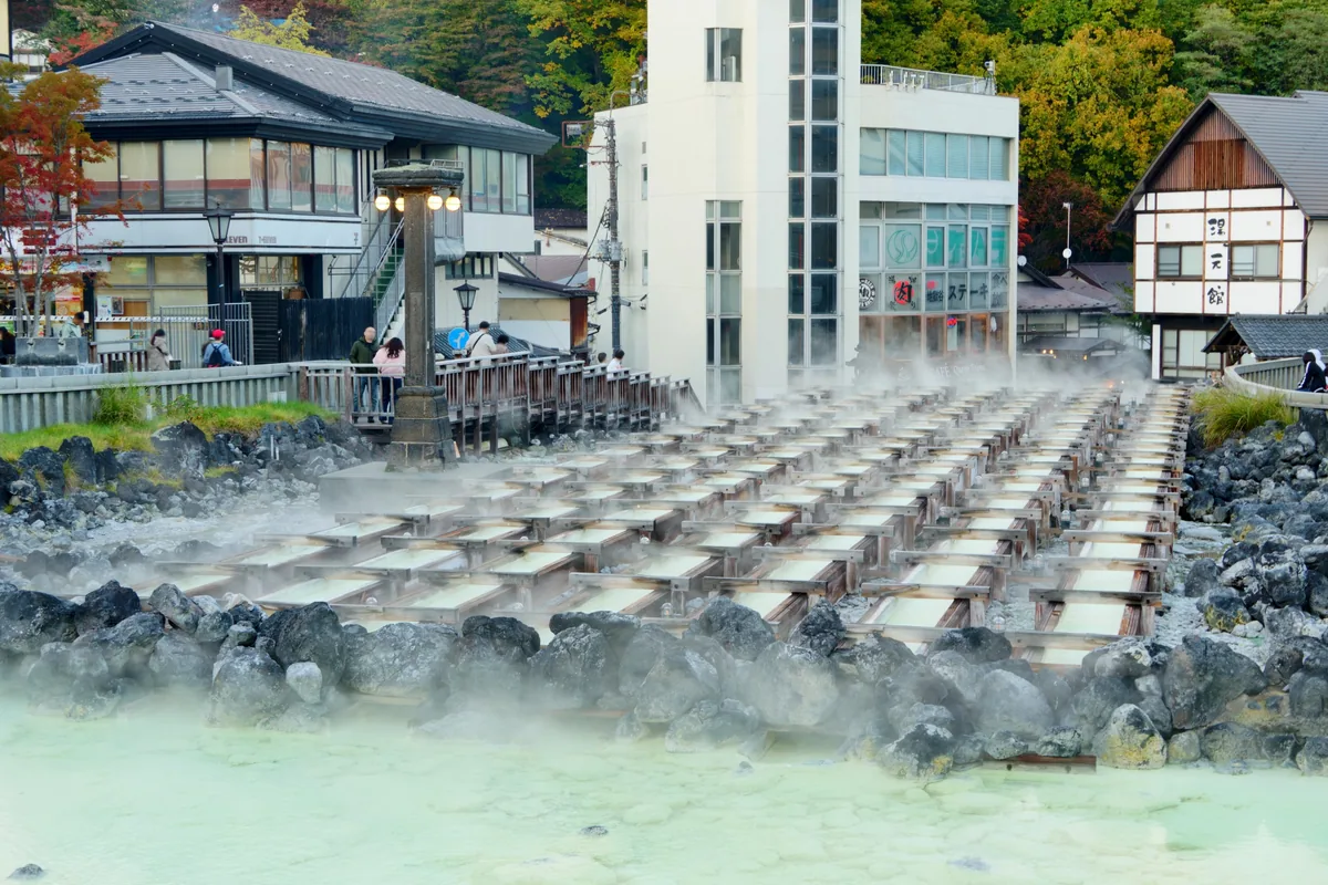 Steam rising from rows of wooden water channels in a hot-spring town, surrounded by buildings and autumn trees