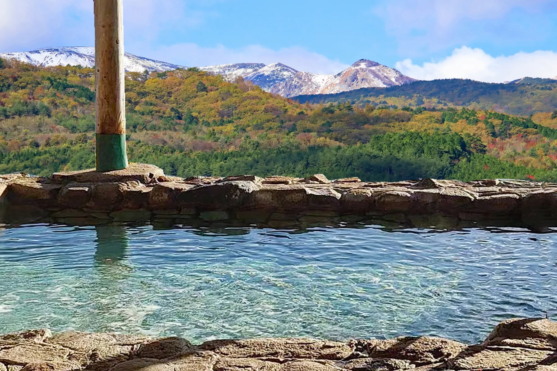 Clear water in a stone-edged outdoor bath overlooking autumn forests and snow-dusted mountain peaks
