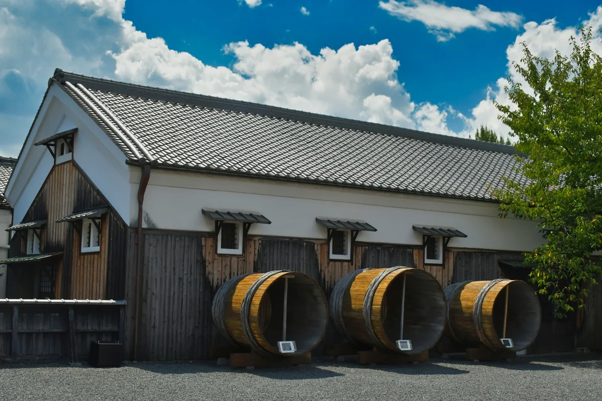 A white-walled, tiled-roof brewhouse with several wooden barrels outside under a blue sky
