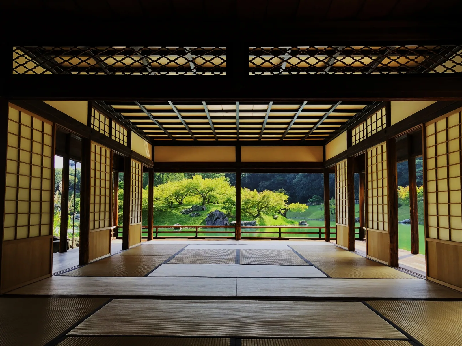 An empty tatami room with open sliding doors framing green trees, rocks and a pond