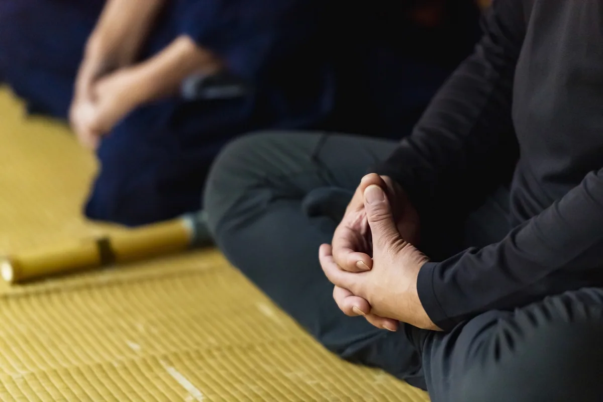 Close view of clasped hands resting in the lap of a person seated on tatami, with another person softly out of focus
