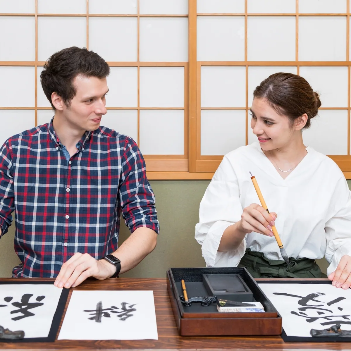 Two visitors holding brushes while practicing Japanese calligraphy indoors