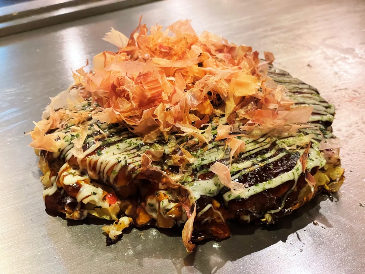 Okonomiyaki topped with sauce, mayonnaise, seaweed and bonito flakes on a metal griddle