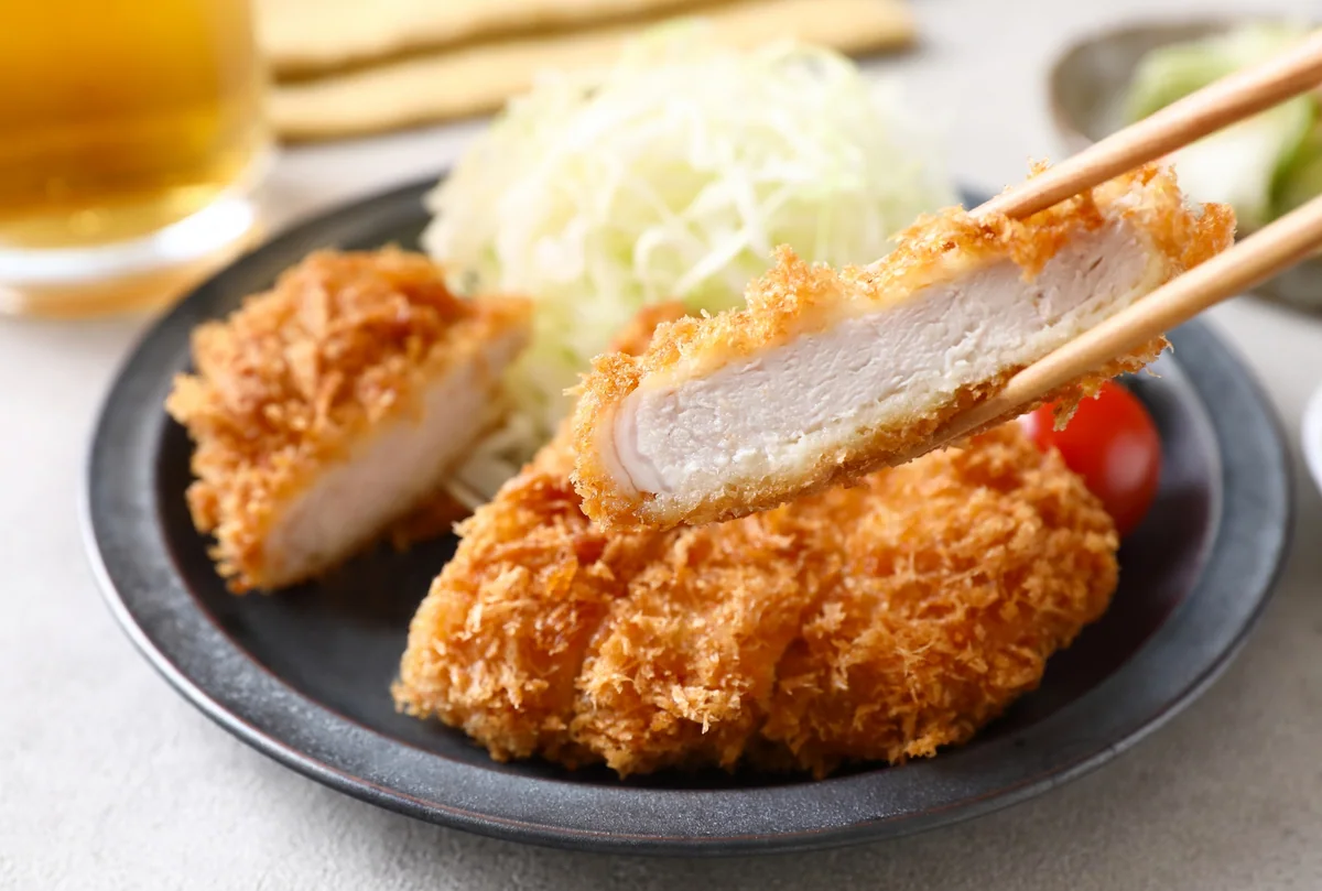 A piece of breaded pork cutlet held with chopsticks above a plate of tonkatsu and shredded cabbage
