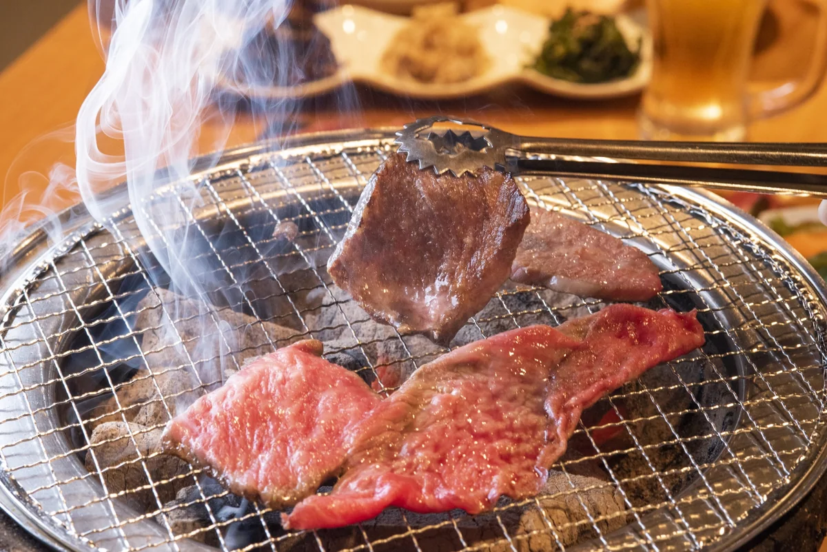 Pieces of meat cooking on a tabletop wire grill, with one piece held in tongs
