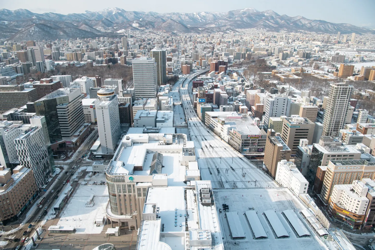 Snow-covered rooftops and railway tracks in Sapporo, with mountains beyond the city
