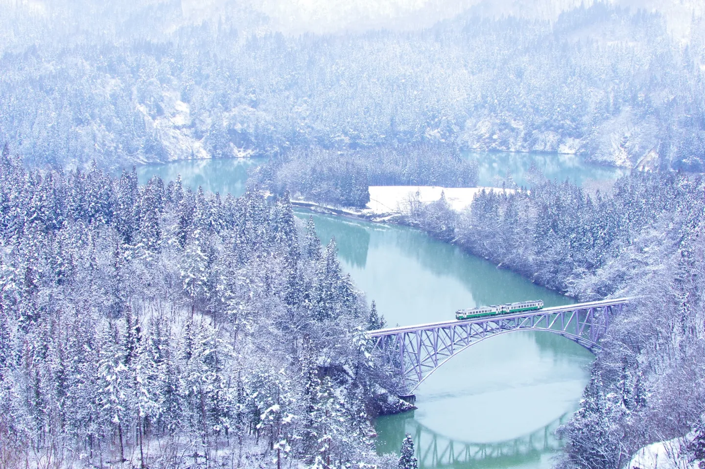 A green-and-white train crossing the First Tadami River Bridge between snowy wooded slopes